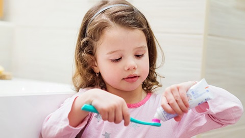 Menina de pijama cor de rosa a pôr dentífrico numa escova azul para lavar os dentes.